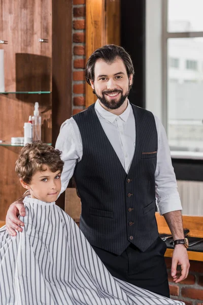 Peluquero barbudo guapo con niño pequeño cubierto con tela rayada mirando a la cámara en la barbería - foto de stock
