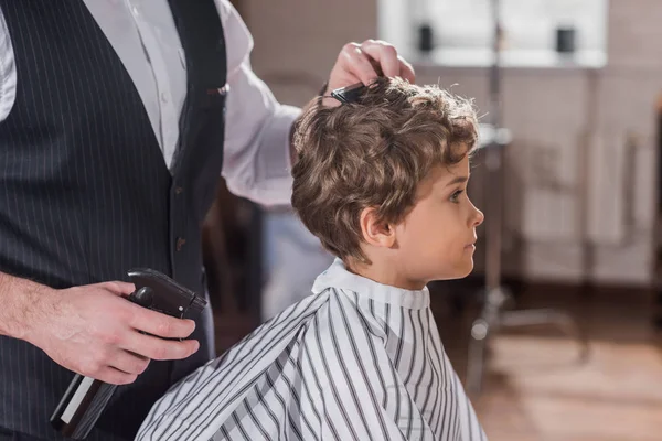 Обрезанный снимок парикмахера, расчесывающего волосы маленького ребенка в детской парикмахерской — стоковое фото