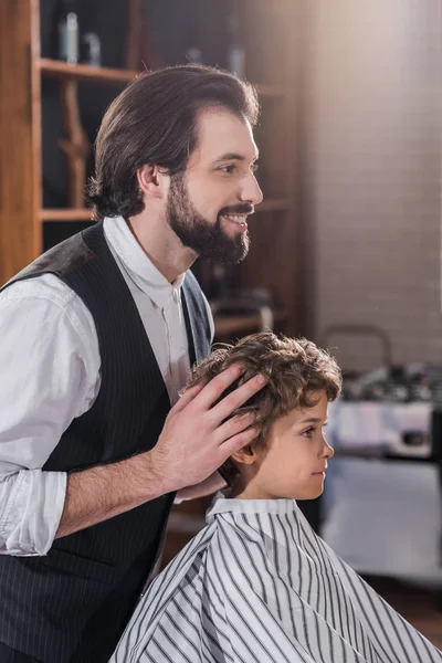 Beau barbier souriant regardant le reflet d'un petit enfant assis sur une chaise au salon de coiffure — Photo de stock