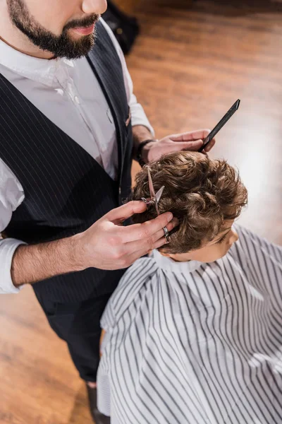 Visão de alto ângulo do garoto encaracolado coberto com pano listrado sentado na barbearia enquanto o barbeiro cortando seu cabelo — Fotografia de Stock