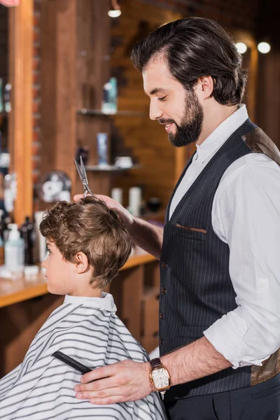 Side view of little curly kid covered with striped cloth sitting at barbershop while barber cutting his hair — Stock Photo