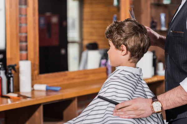 Petit enfant bouclé recouvert de tissu rayé assis au salon de coiffure tandis que le coiffeur couper ses cheveux — Photo de stock