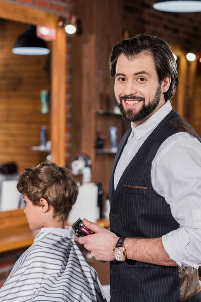 Friseur schneidet Haare eines kleinen Kindes mit Haarschneidemaschine und schaut in die Kamera — Stockfoto