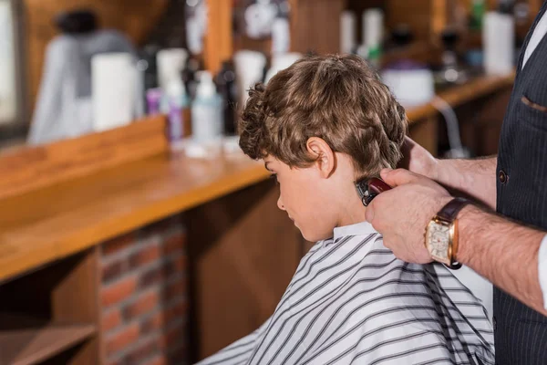 Pequeno garoto encaracolado recebendo corte de cabelo de barbeiro com Hair Clipper — Fotografia de Stock