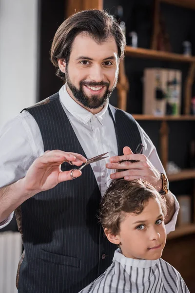 Adorable niñito consiguiendo corte de pelo de guapo sonriente peluquero en niños barbería - foto de stock