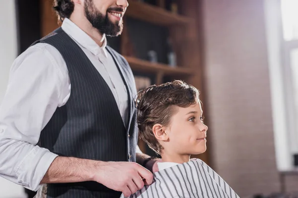 Barbier barbu souriant couvrant adorable petit enfant avec tissu rayé au salon de coiffure pour enfants — Photo de stock