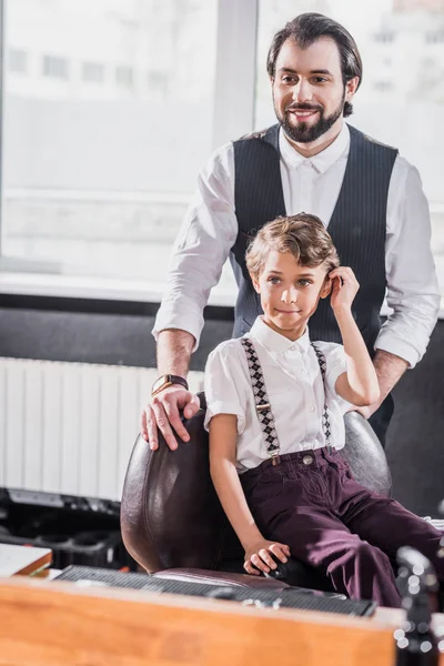 Pequeño niño elegante sentado en la silla en la barbería de los niños con el peluquero en el fondo - foto de stock