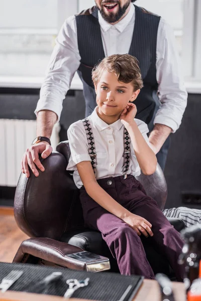 Entzückendes kleines Kind sitzt auf Stuhl beim Kinderfriseur mit Friseur im Hintergrund — Stockfoto