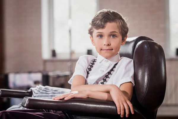 Niño pequeño y elegante sentado en la silla de barbero en la barbería y mirando a la cámara - foto de stock