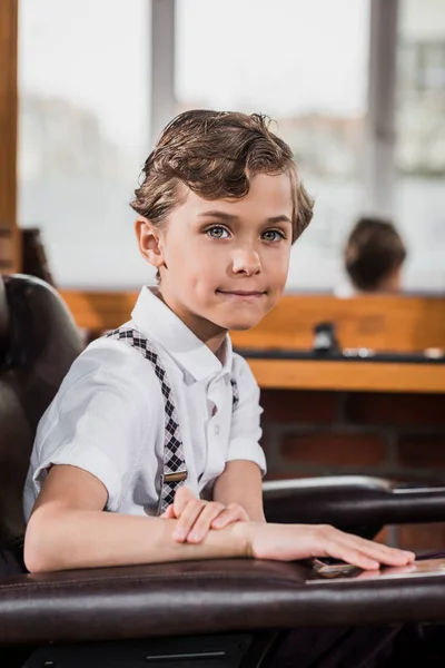 Adorable little kid sitting in barber chair at barbershop and looking at camera — Stock Photo