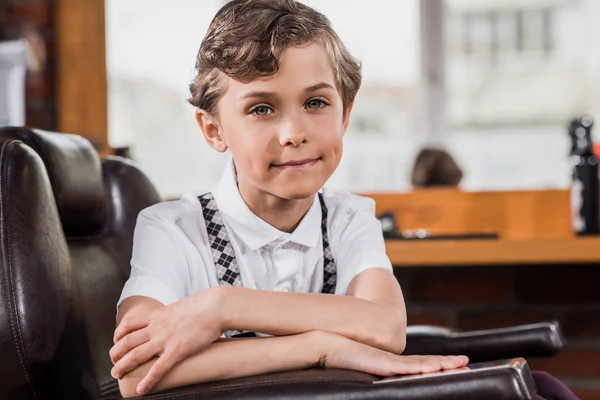 Niño pequeño sentado en silla de barbero en la barbería y mirando a la cámara - foto de stock