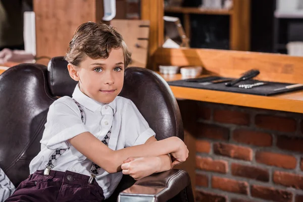 Bambino sorridente seduto sulla sedia del barbiere al barbiere e guardando la macchina fotografica — Foto stock