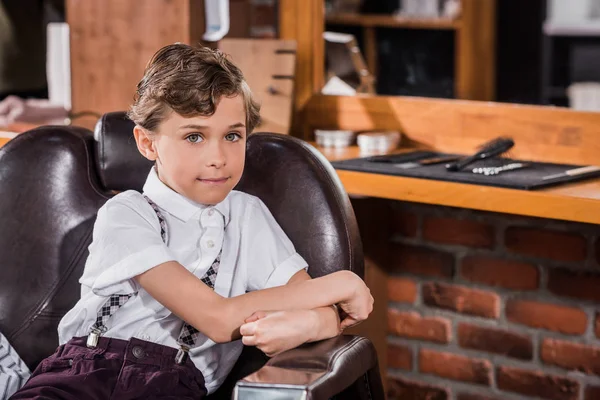 Niño sonriente sentado en la silla de barbero en la barbería y mirando a la cámara - foto de stock