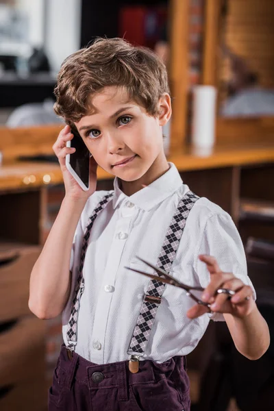Niño pequeño con tijeras hablando por teléfono en la barbería - foto de stock