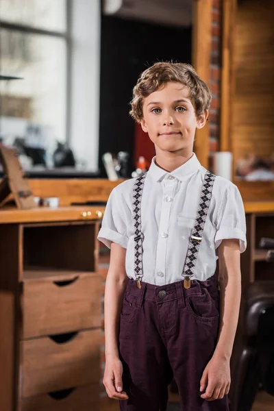 Stylisches kleines Kind im weißen Hemd mit Hosenträgern blickt beim Friseur in die Kamera — Stockfoto