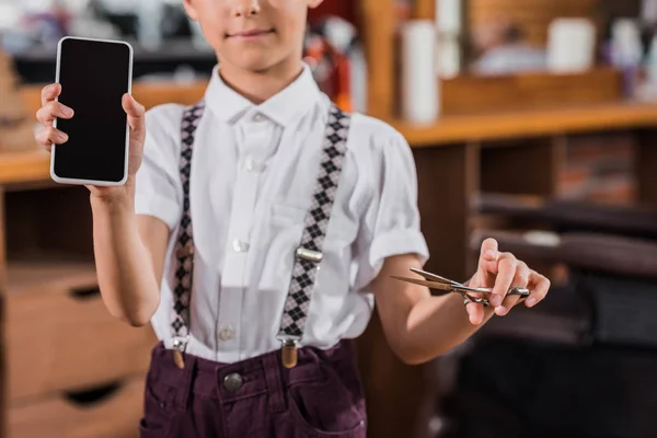 Tiro recortado de niño pequeño con tijeras y teléfono inteligente en la barbería - foto de stock