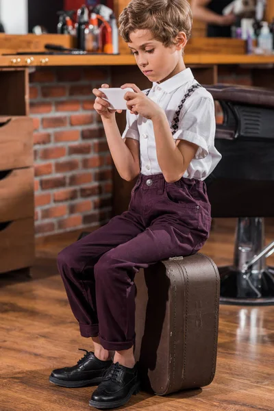 Adorable niño pequeño usando teléfono inteligente mientras está sentado en la maleta en la barbería - foto de stock