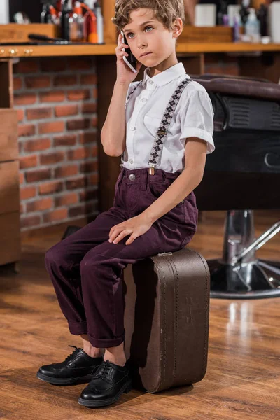 Adorable niño pequeño hablando por teléfono mientras está sentado en la maleta en la barbería - foto de stock