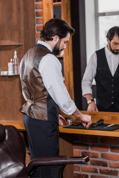 Guapo joven peluquero en elegante chaleco tomando herramientas de estera de goma en el lugar de trabajo - foto de stock