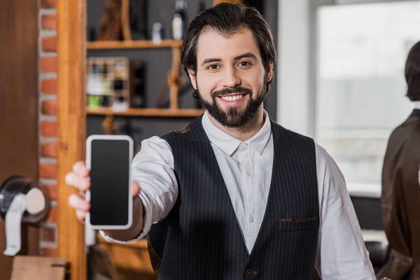 Sonriente joven peluquero en chaleco apuntando al teléfono inteligente - foto de stock