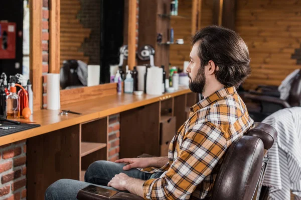 Homem barbudo bonito em camisa xadrez sentado na cadeira na barbearia — Fotografia de Stock