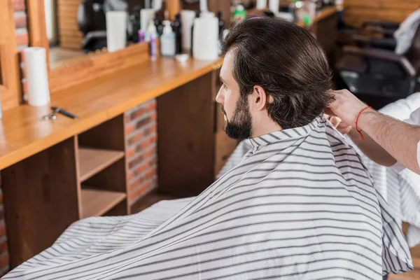 Bearded young man covered with striped cloth sitting on chair at barbershop — Stock Photo