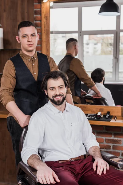 Happy young customer and barber in barbershop looking at camera — Stock Photo