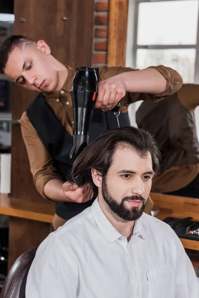 El peluquero serio que peina los cabellos del cliente con el secador de pelo en la barbería - foto de stock