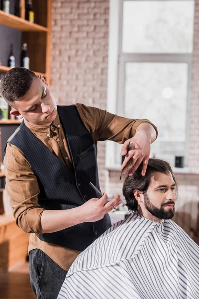 Bearded young man covered with striped cloth getting haircut from professional barber at barbershop — Stock Photo