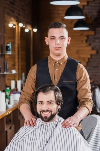 Beau jeune coiffeur debout derrière client sur le lieu de travail — Photo de stock