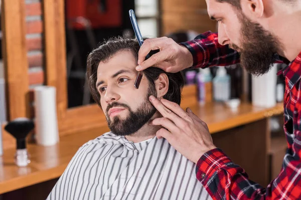 Молодой парикмахер в клетчатой рубашке бреется человек с парикмахерской — стоковое фото