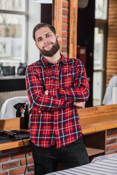 Guapo joven peluquero en camisa a cuadros recostado en el lugar de trabajo en la barbería - foto de stock