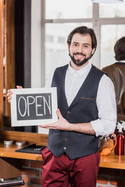 Happy bearded barber holding open signboard in front of workplace — Stock Photo