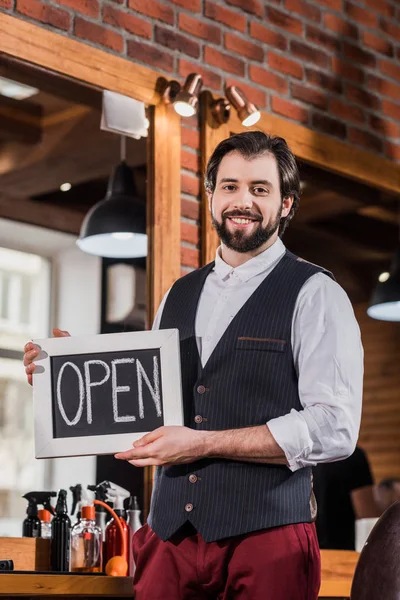 Handsome bearded barber holding open signboard in front of workplace — Stock Photo