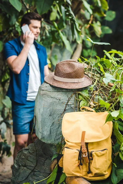 Красивый молодой человек разговаривает по телефону в тропических лесах с рюкзаком и соломенной шляпой на переднем плане — стоковое фото