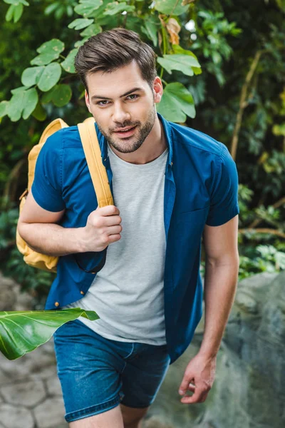 Jeune homme souriant dans des vêtements élégants avec sac à dos marche dans le parc — Photo de stock