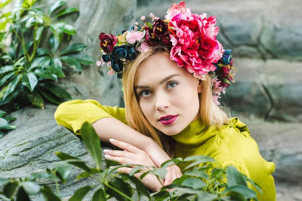 Beautiful young woman with floral wreath leaning on rocks and looking at camera — Stock Photo