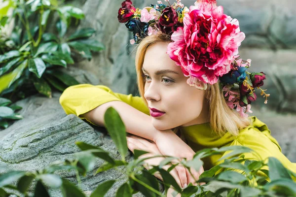 Hermosa joven con corona floral apoyada en rocas y mirando hacia otro lado - foto de stock