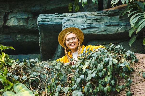 Blick von unten auf glückliche junge Frau mit Strohhut, die im Dschungel in die Kamera schaut — Stockfoto