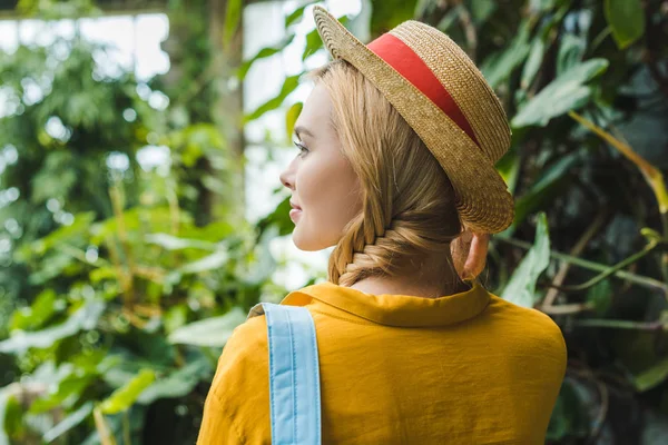 Vue arrière de belle jeune femme en chapeau de paille entourée de plantes tropicales — Photo de stock