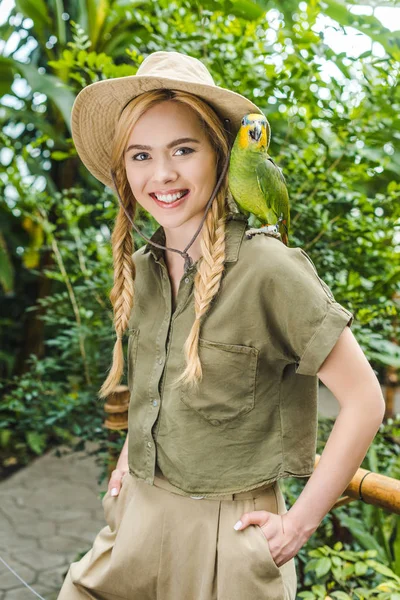 Улыбающаяся молодая женщина в костюме сафари с попугаем на плече в парке джунглей — стоковое фото
