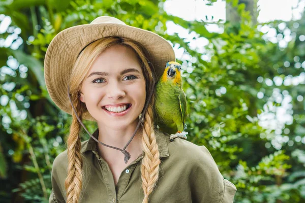 Jeune femme heureuse en costume de safari avec perroquet sur l'épaule dans la jungle — Photo de stock