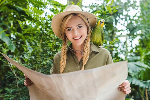 Attraente giovane donna in tuta safari con pappagallo sulla spalla che naviga nella giungla con mappa — Foto stock