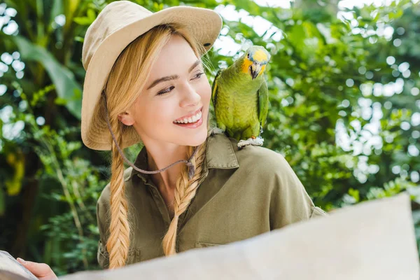 Усміхнена молода жінка в сафарі костюм дивиться на папугу на плечі під час навігації в джунглях з картою — стокове фото