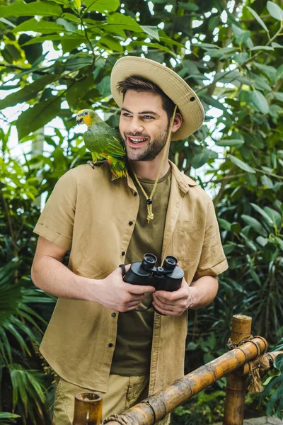 Jovem sorridente com papagaio no ombro e binóculos na selva — Fotografia de Stock