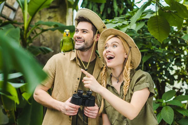 Aufgeregtes junges Paar in Safarianzügen mit Papagei im Regenwald — Stockfoto