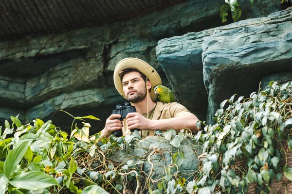 Bottom view of young man with parrot on shoulder and binoculars looking at camera — Stock Photo