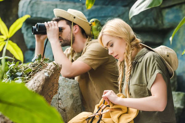 Activa pareja joven en trajes de safari con loro senderismo en la selva - foto de stock