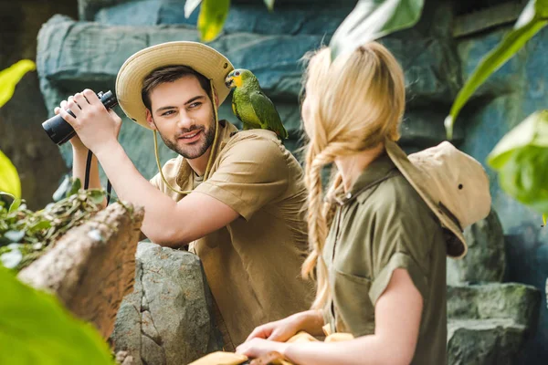 Jovem em terno safari com papagaio no ombro flertando com a mulher enquanto caminhava na selva — Fotografia de Stock