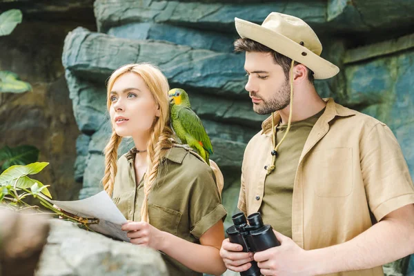 Belo jovem casal em ternos de safári com papagaio tentando navegar na selva — Fotografia de Stock